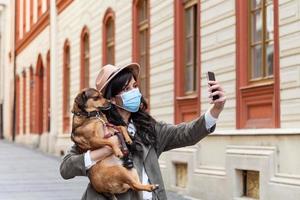 mujer durante el aislamiento pandémico caminando con su perro y usando el teléfono móvil para hacer un selfie. chica elegante con un perro divertido descansando, abrazándose y divirtiéndose, lindos momentos. foto