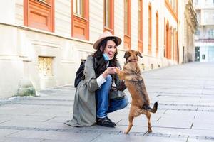 Young woman with long hair and face protective mask embracing funny dog on the street during coronavirus pandemic isolation. Smiling young woman enjoying good day and posing with pet. photo