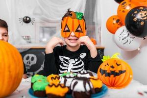 lindo niño con máscara de calabaza disfrutando de su halloween. Jack o' Lantern calabaza de Halloween en la mesa y otras decoraciones de miedo foto