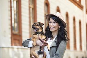 Young smiling woman with a hat in casual clothes iron and hugging dog whilewalking on the street. Smiling young woman enjoying good day and posing with pet. Woman playing with her dog photo