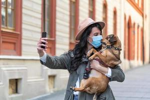 Woman during pandemic isolation walking with her dog and using mobile phone to make a selfie. Stylish girl with funny dog resting, hugging and having fun, cute moments. photo