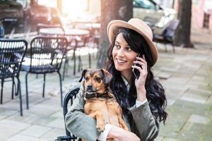 Attractive young woman sitting in coffee shop, relaxing and holding her dog while talking on the phone. Pet friendly cafeteria, beautiful girl with her dog siting at a coffee shop and drink coffee. photo