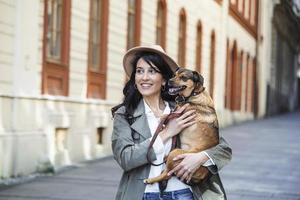 Young smiling woman with a hat in casual clothes iron and hugging dog whilewalking on the street. Smiling young woman enjoying good day and posing with pet. Woman playing with her dog photo