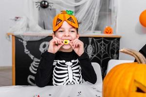 lindo niño con disfraz de miedo disfrutando de sus dulces de halloween. Jack o' Lantern calabaza de Halloween en la mesa y otras decoraciones de miedo foto