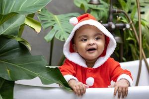 Portrait of African American little toddler baby girl in santa christmas dress smiling and playing inside the bathtub surrounding by plant photo