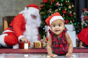 el bebé afroamericano se divierte jugando con juguetes mientras santa claus prepara un regalo y se sienta detrás del árbol de navidad para el concepto de celebración de temporada foto