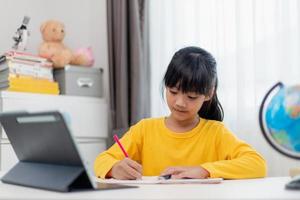 Asian schoolgirl doing her homework with digital tablet at home. Children use gadgets to study. Education and distance learning for kids. Homeschooling during quarantine. Stay at home photo