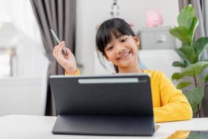 Asian schoolgirl doing her homework with digital tablet at home. Children use gadgets to study. Education and distance learning for kids. Homeschooling during quarantine. Stay at home photo