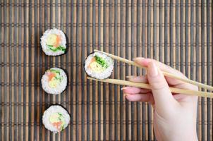 A hand with chopsticks holds a sushi roll on a bamboo straw serwing mat background. Traditional Asian food photo