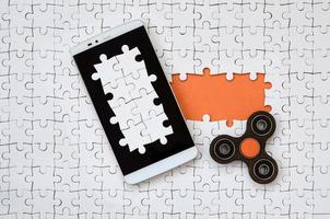 A modern big smartphone with a touch screen and a spinner lie on a white jigsaw puzzle in an assembled state with missing elements photo