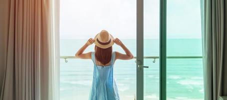 Happy woman wearing blue dress and hat looking outside window to ocean view in morning. Relax, vacation, time to travel and freedom concept photo