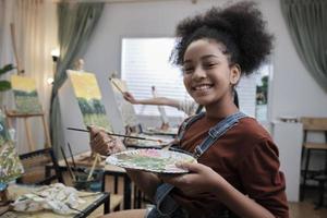 One Black student girl smiling with paintbrush and palette, acrylic color picture painting on canvas in art classroom and creative learning with talents skill at elementary school studio education. photo