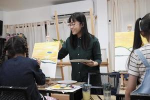 A female Asian teacher teaches and demonstrates to the children on acrylic color picture painting on canvas in art classroom, creatively learning with skill at the elementary school studio education. photo