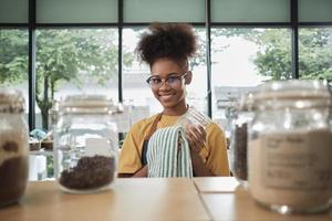 una joven comerciante negra limpia frascos de vidrio de productos orgánicos naturales en contenedores reutilizables en una tienda de recarga, cero desperdicio, una tienda de comestibles sin plástico y un negocio minorista ecológico. foto