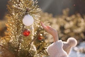 Child girl is decorating Christmas tree with shiny white and red balls outdoors before the holidays. Kid in warm jacket is having fun in park in winter day. Merry Xmas and happy family holidays photo