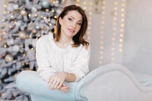 A young, attractive woman in a white sweater sits on the sofa near the Christmas tree and light photo