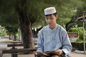 Young asian muslim boy wears hat, sitting in school park and reading his book in his free times before going back home, soft and selective focus. photo