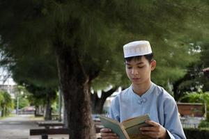 Young asian muslim boy wears hat, sitting in school park and reading his book in his free times before going back home, soft and selective focus. photo