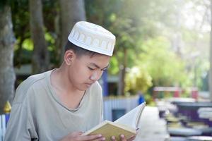 Young asian muslim boy wears hat, sitting in school park and reading his book in his free times before going back home, soft and selective focus. photo