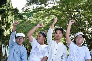 Young asian Muslim boys are raising their right hands to show their victory and success happily under a tree in the school park, soft and selective focus. photo