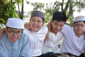 Young asian Muslim boys are playing with their hands behind each other by sitting in a row happily under a tree in the school park, soft and selective focus. photo