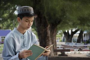 Young asian muslim boy wears hat, sitting in school park and reading his book in his free times before going back home, soft and selective focus. photo