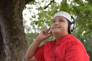 Portrait Young asian muslim boy in brown shirt and wears hat, holds headphone and sitting under the tree in the park, soft and selective focus. photo