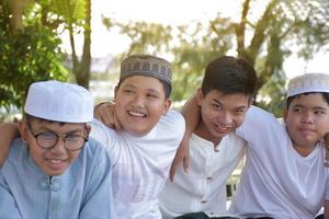 Young asian Muslim boys are playing with their hands behind each other by sitting in a row happily under a tree in the school park, soft and selective focus. photo