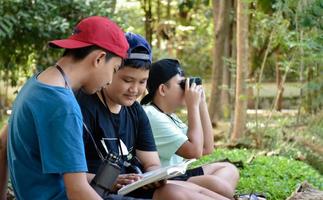 Three asian boys're reading birds details and going to use binoculars to watch birds on the trees during summer camp, idea for learning creatures and wildlife animals outside the classroom. photo