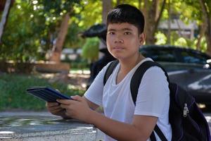 Portrait young asian boy holds laplet and stands under the big tree, soft focus, concept for using technology in daily life. photo