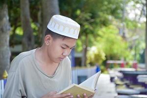 Young asian muslim boy wears hat, sitting in school park and reading his book in his free times before going back home, soft and selective focus. photo