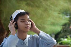 retrata a un joven musulmán asiático con camisa marrón y usa sombrero, sostiene auriculares y se sienta bajo el árbol en el parque, enfoque suave y selectivo. foto