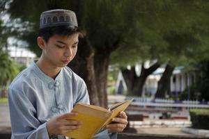 Young asian muslim boy wears hat, sitting in school park and reading his book in his free times before going back home, soft and selective focus. photo