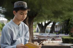 Young asian muslim boy wears hat, sitting in school park and reading his book in his free times before going back home, soft and selective focus. photo