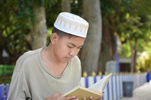 Young asian muslim boy wears hat, sitting in school park and reading his book in his free times before going back home, soft and selective focus. photo