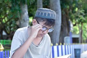 Young asian muslim boy wears eyeglasses, sitting in school park and reading his book in his free times before going back home, soft and selective focus. photo