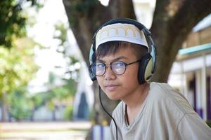 Portrait Young asian muslim boy in brown shirt and wears hat, holds headphone and sitting under the tree in the park, soft and selective focus. photo