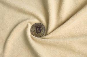 The golden bitcoin lies on a blanket made of soft and fluffy light orange fleece fabric with a large number of relief folds. The shape of the folds resembles a fan from a video card cooler photo