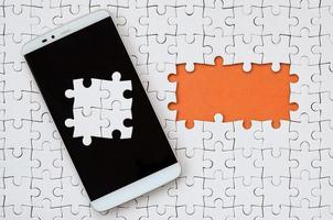 A modern big smartphone with several puzzle elements on the touch screen lies on a white jigsaw puzzle in an assembled state with missing elements photo