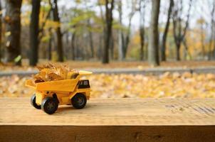 The concept of seasonal harvesting of autumn fallen leaves is depicted in the form of a toy yellow truck loaded with leaves against the background of the autumn park photo