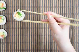 A hand with chopsticks holds a sushi roll on a bamboo straw serwing mat background. Traditional Asian food photo