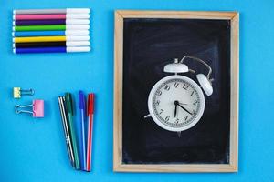 pizarra escolar y accesorios de papelería, lápices, reloj despertador y bolígrafos sobre fondo azul. concepto de regreso a la escuela. foto