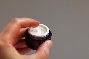 Hands of a girl with a jar of face cream close up applied to the finger of woman's hand on light background photo