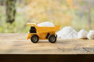 A small yellow toy truck is loaded with a stone of white salt next to a pile of salt. A car on a wooden surface against a background of autumn forest. Extraction and transportation of salt photo