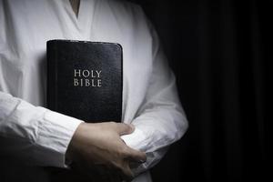 Faith with holy bible concept. Hands of a female prayer worship God with holy bible on black background in church. Christian woman who believe in Jesus read and study the grace of the holy scriptures.