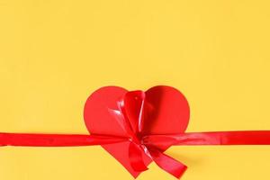 Red ribbon with bow and heart on a yellow background, top view. Flat lay, copy space. photo