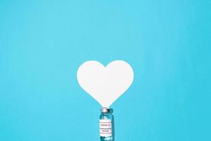 Ampoule of COVID-19 vaccine and white heart on a blue background, flat lay. Top view, copy space. Global population vaccination, healthcare and medicine concept photo