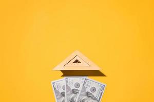 Wooden roof of toy house and one hundred dollar bills on yellow background, flat lay. Concept of buying a home, real estate cost, renting an apartment, investing in construction. Top view, copy space photo