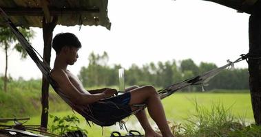 un joven granjero sin camisa se sienta en la hamaca, escribe en una computadora portátil y mira los campos de arroz, agita el sombrero en la mano para refrescarse video
