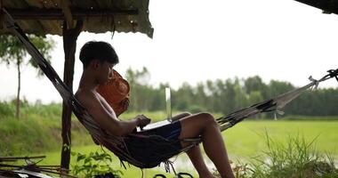vista lateral de un joven granjero sin camisa sentado en la hamaca y mirando los campos de arroz, agitando el sombrero en la mano para refrescarse video